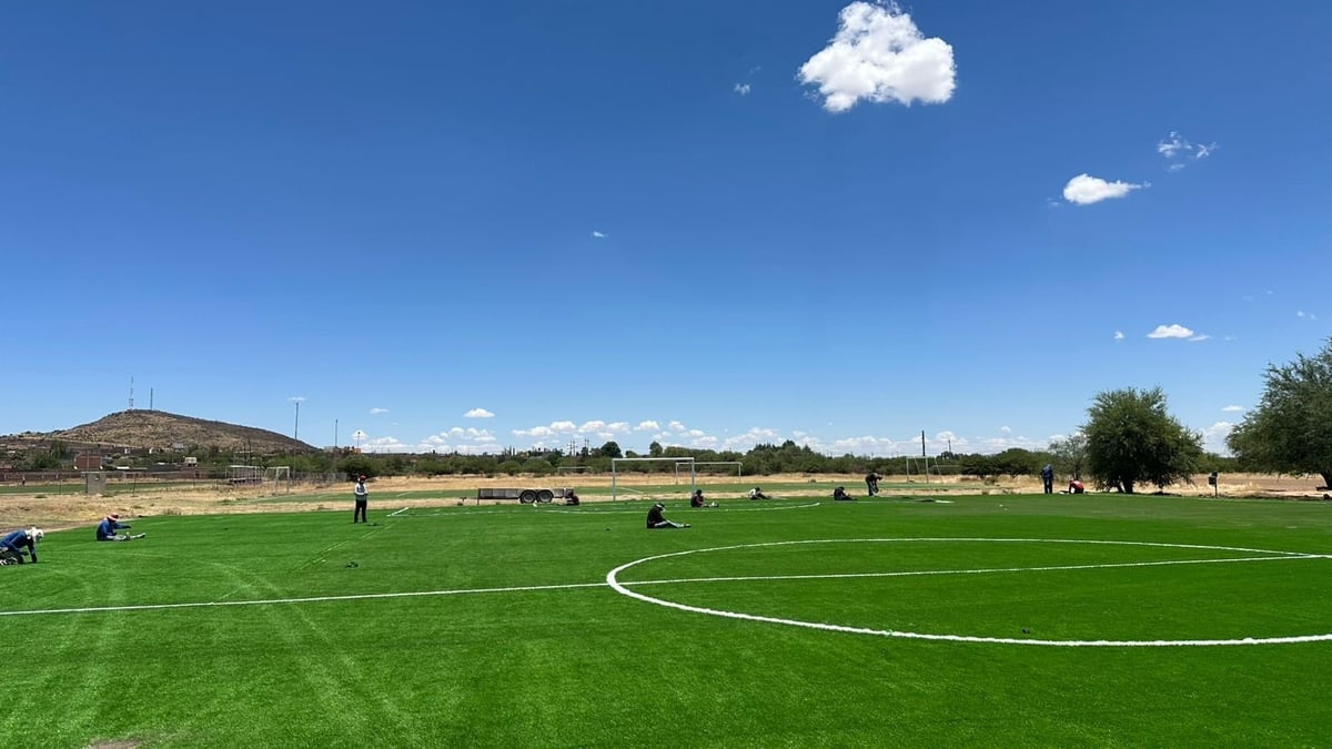 Amplia vista de campo de fútbol con jugadores, líneas blancas y montañas al fondo bajo cielo azul